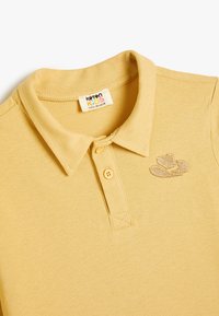 Keltainen polo-paita, joka on valmistettu teksturoidusta puuvillasta. Siinä on kaulus, kaksi nappia ja pieni brodeerattu hattu-logo vasemmassa rinnassa.