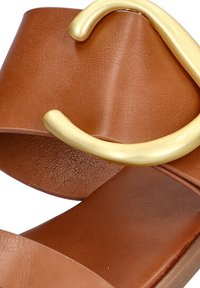 Sandal de cuero marrón con una tira ancha y un adorno metálico dorado, suela plana y textura suave.