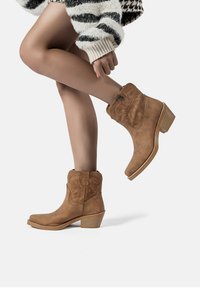 Stivali ankle in suede beige con cuciture decorative e tacco impilato. La silhouette è slanciata, con punta tonda e un dettaglio con bottone laterale.