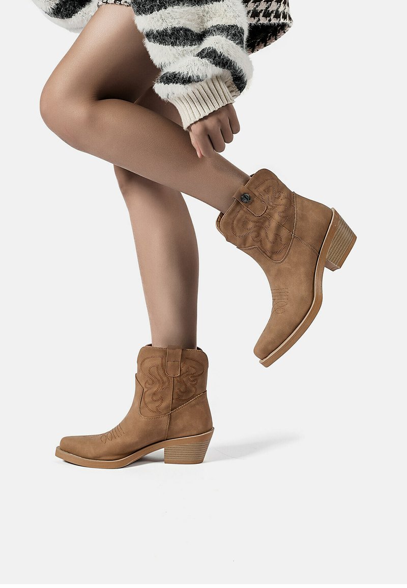 Stivali ankle in suede beige con cuciture decorative e tacco impilato. La silhouette è slanciata, con punta tonda e un dettaglio con bottone laterale.