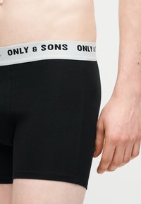 Αρσενικός κορμός και χέρι που φοράει μαύρα σλιπ μποξέρ με λευκή ζώνη μέσης με την ένδειξη «ONLY & SONS» σε απλό φωτεινό φόντο.