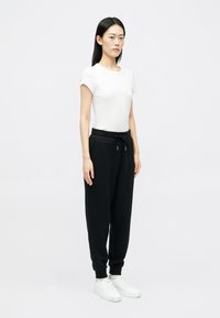 Jauna sieviete stāv, ģērbusies pielāgotā baltā īsās piedurknes topā, melnos sporta biksēs ar auklu un baltos sporta apavos, uz neitrāla fona.