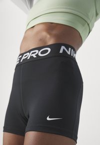 Gros plan sur une personne portant un short de compression Nike Pro noir avec un logo visible et un haut court vert clair.