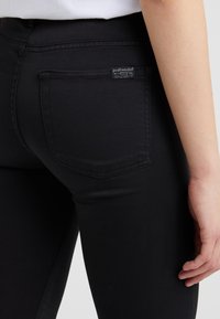 Lähiotokuva henkilöstä, joka käyttää tiukkoja mustia housuja, joissa on takatasku ja pieni kangasmerkki, käsi lepääntyneenä reiden vieressä.