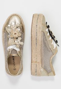 Zapatillas de malla dorada con adornos florales, suelas de esparto y detalles texturizados. Presentan cordones y acentos decorativos en blanco y negro.