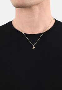 Collar de cadena de oro con un pequeño colgante de letra "R" texturizada, llevado sobre una camiseta negra, con un diseño de cadena fina y entrelazada.