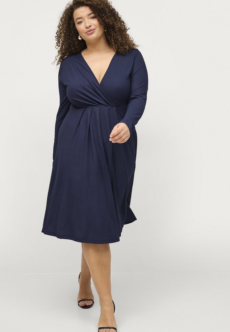 Marineblaues Wickelkleid aus strukturiertem Stoff, V-Ausschnitt, lange Ärmel und fließender Saum. Tragen mit schwarzen Riemchenheels.
