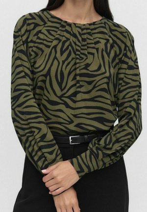 Kobieta w zielono-czarnej, zebrowanej bluzce z długim rękawem, z plisami przy dekolcie i czarnym paskiem do czarnych spodni.