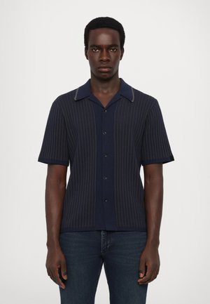 Navy short-sleeve shirt with vertical stripes, button-up front, contrasting collar, and textured fabric. Worn with dark jeans.