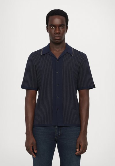 Chemise bleu marine à manches courtes avec des rayures verticales, boutonnée à l'avant, col contrasté et tissu texturé. Portée avec un jean foncé.