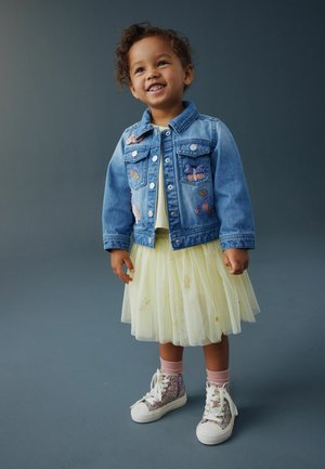 Uśmiechnięte dziecko w niebieskiej dżinsowej kurtce z naszywkami, żółtej spódniczce z tiulu, różowych skarpetkach i błyszczących trampkach na wysokiej podeszwie, stojące na szarej podłodze.