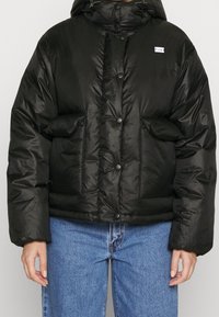 Sort pufferjakke med hætte, der har en skinnende overflade, store forlommer og knaplukning, parret med blå denimbukser.