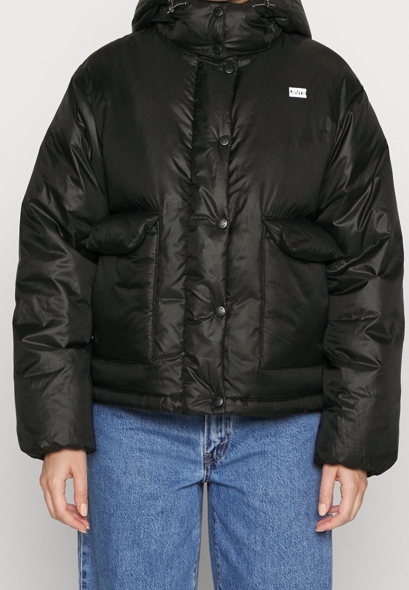Sort pufferjakke med hætte, der har en skinnende overflade, store forlommer og knaplukning, parret med blå denimbukser.