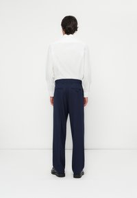 Chemise blanche à manches longues associée à un pantalon bleu marine, avec une coupe droite, des poches arrière et des chaussures noires, sur fond blanc uni.