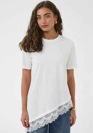 Femme aux cheveux bruns ondulés portant des boucles d'oreilles en or, un haut asymétrique à manches courtes blanc avec un ourlet en dentelle, un jean bleu et un bracelet jonc en or.