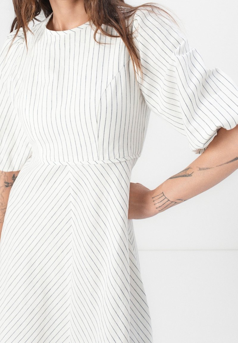 Femme portant une robe blanche avec de fines rayures verticales noires, des manches bouffantes et des tatouages sur l'avant-bras gauche.