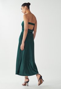 Abito maxi in raso verde con design senza spalline, schiena arricciata e dettagli in tessuto drappeggiato, abbinato a scarpe con tacco alto nere e con cinturini.