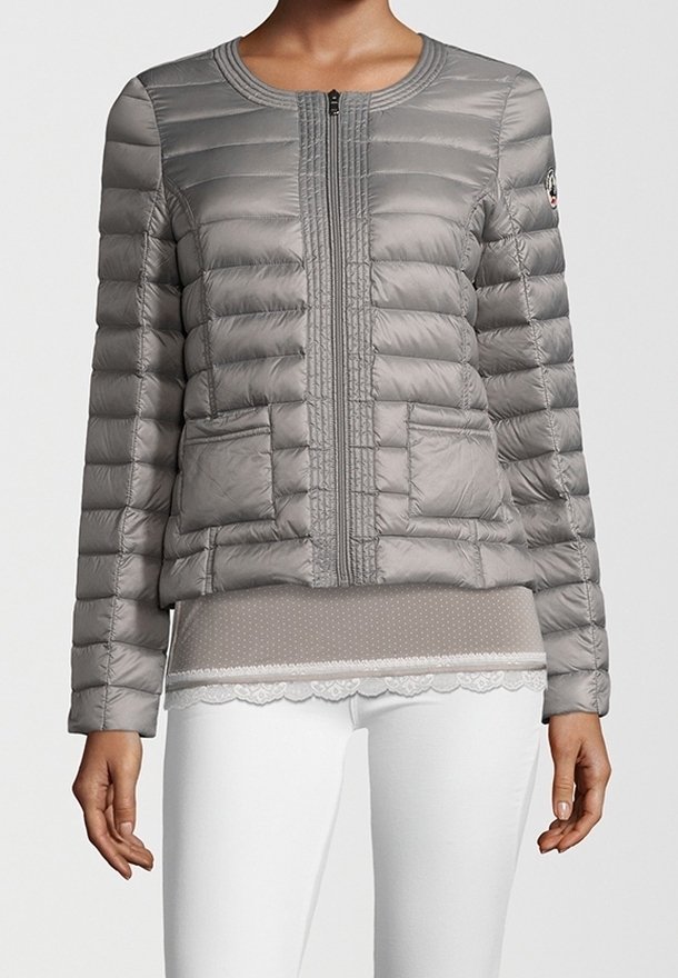 Veste matelassée grise pour femme avec fermeture éclair, manches longues, deux poches avant, portée avec un haut taupe orné de dentelle et un pantalon blanc.