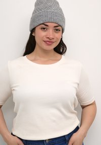 Krémový krátký rukáv triko s kulatým výstřihem a hladkou texturou, doplněné šedou pletenou čepicí. Modelka stojí s rukama na bocích.