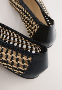 Chaussures plates pour femmes avec talons en cuir noir et corps en rotin tressé noir et beige, vues de dos sur une surface blanche.