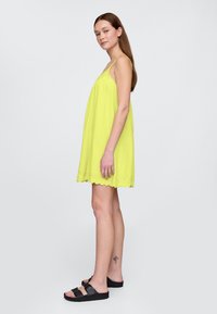 Obleka brez rokavov v limetini zeleni barvi z valovitim robom in vezenim detajlom; kombinirana s črnimi natikači, modelka stoji v stranski pozi.