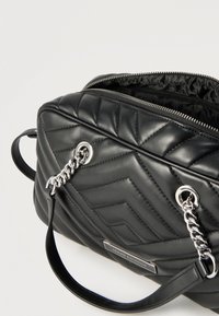 Μαύρη δερμάτινη τσάντα ώμου με καπιτονέ σχέδιο, ασημένιες αλυσίδες και μερικώς ανοιχτό φερμουάρ που αποκαλύπτει την εσωτερική επένδυση με το εμπορικό σήμα.