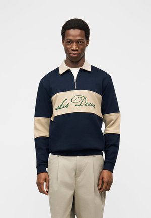 Mladenič v mornarsko modri in bež majici s kapuco z vezenim napisom "Les Deux" na prsih, oblečen v svetlo bež hlače, stoji pred belim ozadjem.