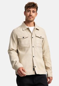 Beige Canvasjacke mit durchgehender Knopfleiste, zwei Brusttaschen, schwarzen Knöpfen und einem lockeren Schnitt. Über einem weißen T-Shirt getragen.
