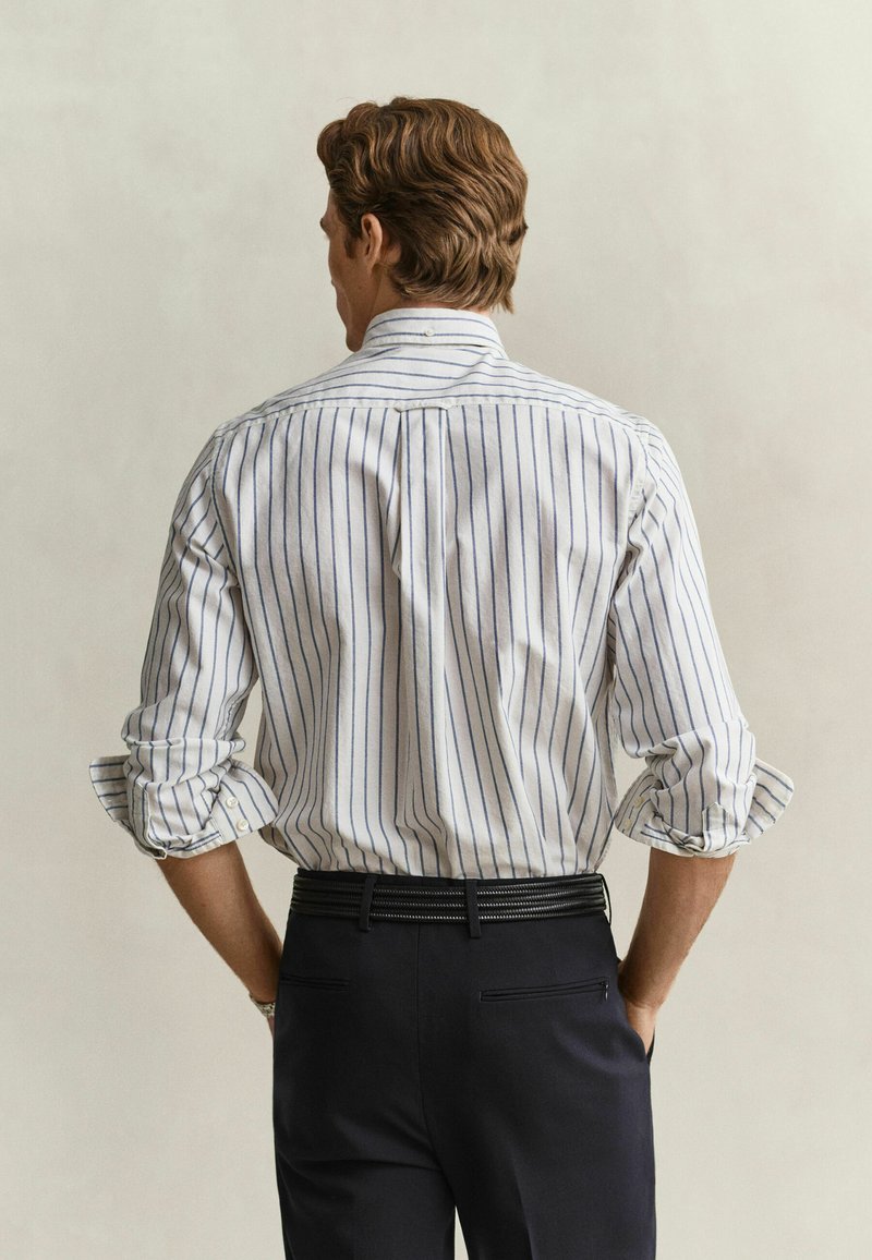 Homme portant une chemise blanche à rayures bleues verticales et un pantalon noir, debout les mains dans les poches, vu de dos sur un fond uni.