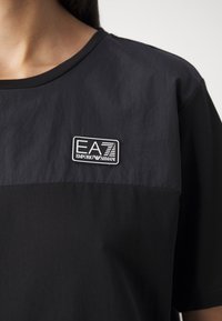 Svart t-shirt med kort ärm som har en kontrasterande tygpanel och en EA7-logotyp. Slät yta med rund halsringning.