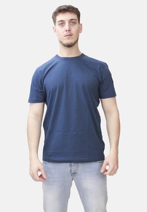 Un giovane uomo è in piedi di fronte, indossando una semplice maglietta blu scuro e jeans blu chiaro su uno sfondo bianco.