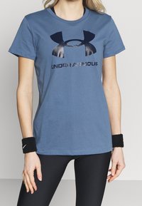 Modré bavlněné tričko s černým logem Under Armour a nápisem. Krátké rukávy a kulatý výstřih. Nosí se s černými legínami a náramky.