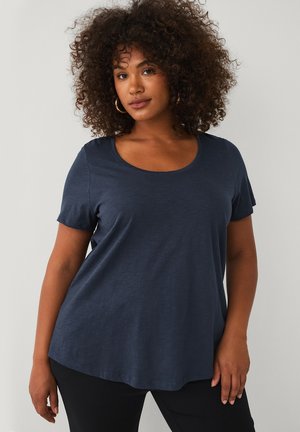 Vrouw met krullend haar die gouden oorringen draagt en een losse donkerblauwe korte mouw blouse, staand tegen een effen achtergrond.