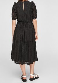 Robe en dentelle noire avec des manches bouffantes, jupe à volants, taille élastique et fermeture par boutons au dos. Associée à des sandales blanches.