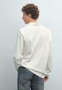 Hvid langærmet polo skjorte med en blød tekstur, med krave. Båret med slidte blå jeans, vist fra ryggen mod en neutral baggrund.