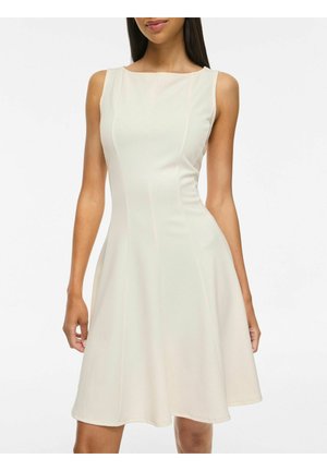 Mujer vestida con un vestido sin mangas, hasta la rodilla, de color beige claro, con un corpiño ajustado y falda acampanada, de pie frente a un fondo blanco liso.