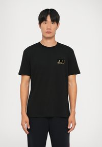 Černé bavlněné tričko s krátkými rukávy, na lev�é straně hrudi se zlatým logem. Klasický kulatý výstřih, hladká textura. Jednoduchý design.