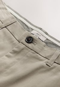 Prim-plan cu pantaloni bej slim fit, cu nasture negru, bucle pentru curea și o etichetă interioară albă în talie cu inscripția "NEXT SLIM FIT."