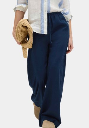 Personne portant un pantalon ample bleu marine et des chaussures beiges, tenant un sac tissé de couleur tan, avec une chemise à rayures claires partiellement rentrée.