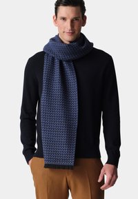 Gestrickter Schal in Blautönen mit strukturiertem Muster und Fransenenden. Über einem dunklen Pullover getragen, zeigt er Wärme und Detailreichtum.