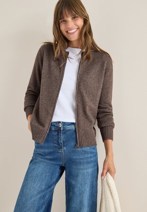 Cardigan marrone con zip anteriore, orlo e polsini a coste. Indossato su una camicia bianca e abbinato a jeans a gamba larga azzurri. Texture morbida.