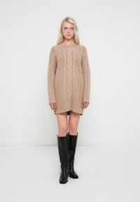 Beige knitted sweater dress with cable knit pattern, long sleeves, and a rounded neckline. Paired with knee-high black leather boots.