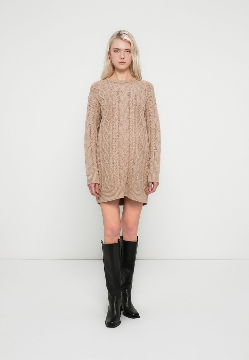 Beige knitted sweater dress with cable knit pattern, long sleeves, and a rounded neckline. Paired with knee-high black leather boots.