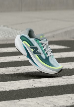 Zapatilla de running New Balance en azul claro y verde con suela blanca, mostrada en el aire sobre un paso de peatones de cebra en un pavimento de asfalto.