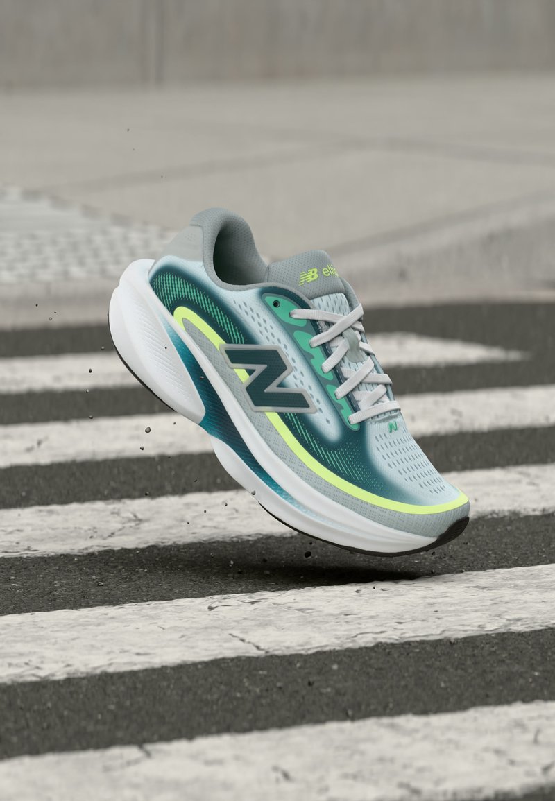 Ανοιχτό μπλε και πράσινο αθλητικό παπούτσι New Balance με λευκή σόλα, εμφανίζεται στον αέρα πάνω από διαβάση ζέβρας σε ασφαλτοστρωμένο πεζοδρόμιο.