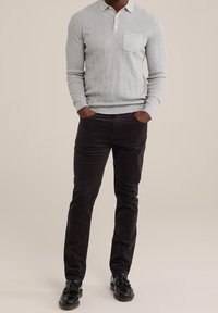 Muž v lehce šedém pleteném polokošili s dlouhým rukávem, tmavě hnědých kalhotách a černých mokasínách s třásněmi, stojící s rukama v kapsách na jednobarevném pozadí.