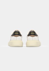 Sneaker bianche con tomaia in pelle liscia e suola spessa color crema. Tassello posteriore nero con logo dorato "BOSS" e accento marrone vicino al tallone.