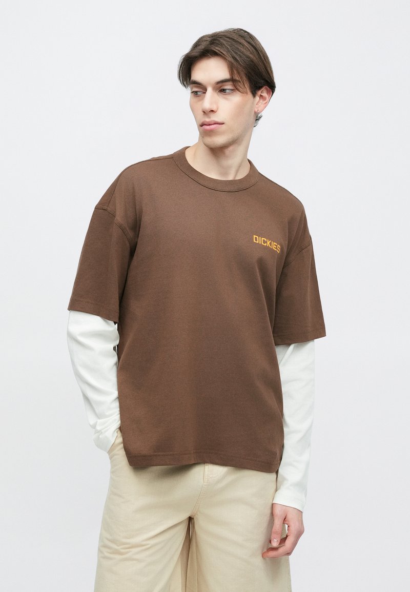 Jeune homme portant un t-shirt marron Dickies par-dessus un sous-pull blanc à manches longues, pantalon beige, regardant vers la gauche, les mains dans les poches, sur un fond uni.
