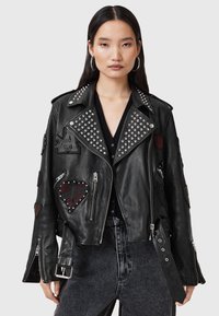 Woman wearing a black studded leather jacket with heart patches and zippers over a black top and high-waisted black jeans.