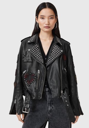 Woman wearing a black studded leather jacket with heart patches and zippers over a black top and high-waisted black jeans.